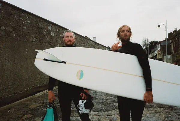 Deux surfeurs en combinaison remontent la cale, l'un portant une planche blanche, l'autre un caisson photo.
