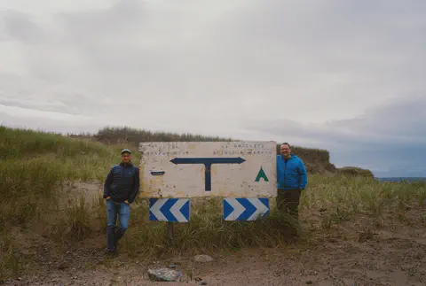 Deux hommes adossés à un vieux panneau de bois dans les dunes, sous un ciel gris.