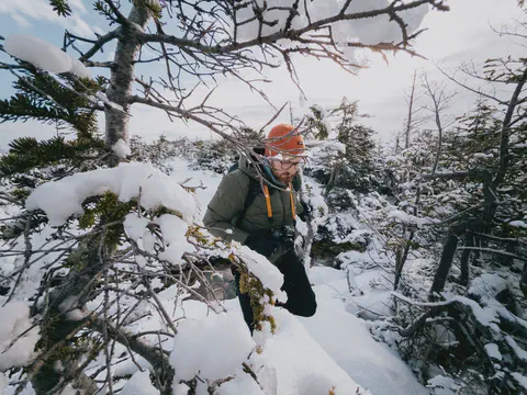 Un photographe progresse entre des sapins chargés de neige, appareil autour du cou.