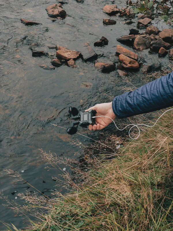 Une main tend un enregistreur et ses micros au-dessus d'un ruisseau bordé de pierres rouges.