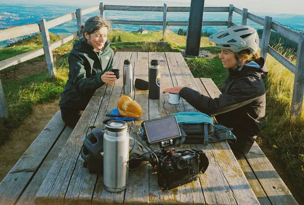 Deux femmes assises à une table de pique-nique en montagne, thermos et caméra posés devant elles, la vallée en contrebas.