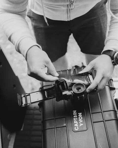 Deux mains engagent un film argentique dans un dos moyen format, posé sur une valise de transport marquée Captain Yvon.
