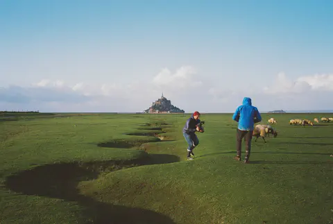 Un opérateur accroupi filme un troupeau de moutons dans les herbus, le Mont-Saint-Michel à l'horizon.