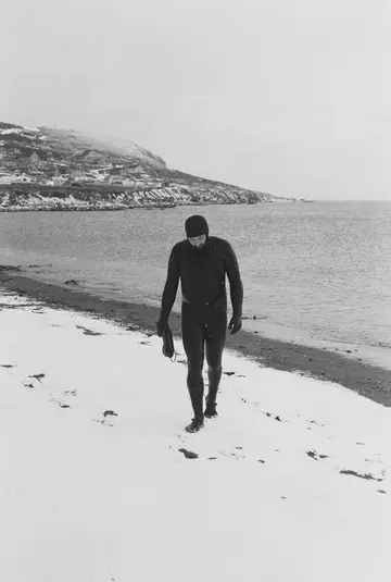 Un surfeur en combinaison remonte une plage recouverte de neige, le village et la falaise derrière lui.
