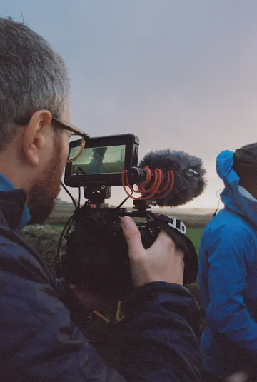 Un opérateur cadre au moniteur déporté, micro monté sur la caméra, dans la lumière rose de fin de journée.