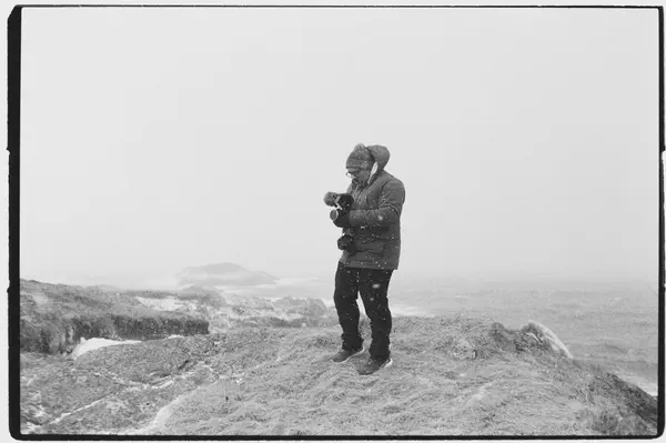 Un homme filme debout sur une pointe rocheuse balayée par la neige, la mer blanche derrière lui.