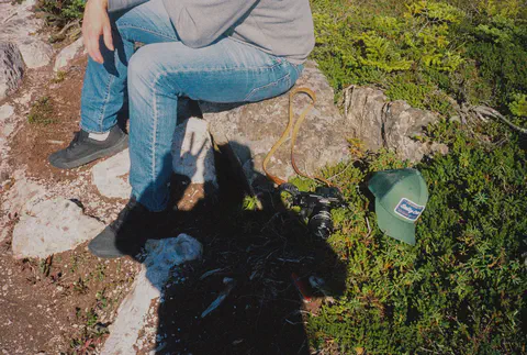 Un photographe assis sur un rocher, son appareil et sa casquette posés dans la végétation rase.